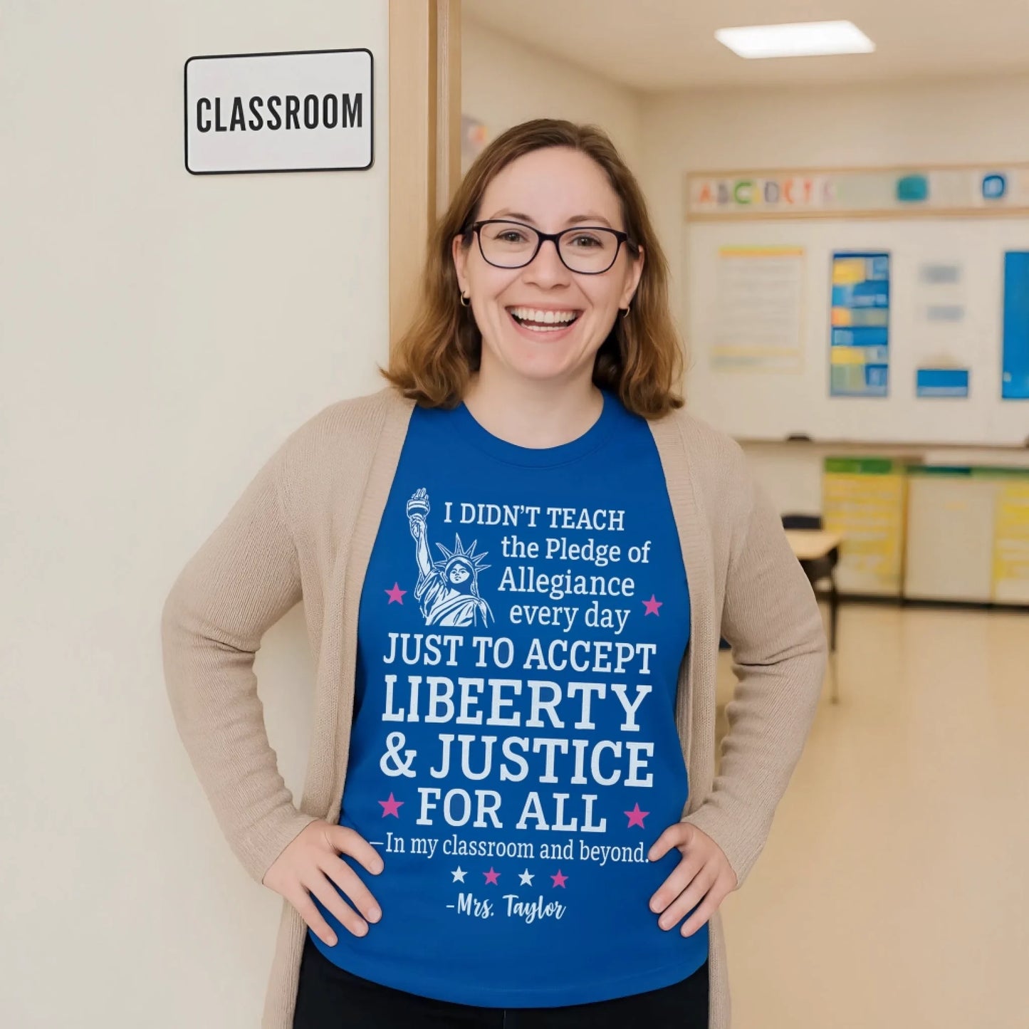 Smiling female teacher wearing a royal blue patriotic T-shirt stands proudly at the doorway of a classroom with a classroom sign beside her and educational posters visible in the bright organized room behind her