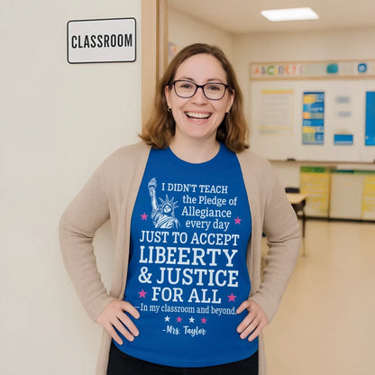 Smiling female teacher wearing a royal blue patriotic T-shirt stands proudly at the doorway of a classroom with a classroom sign beside her and educational posters visible in the bright organized room behind her